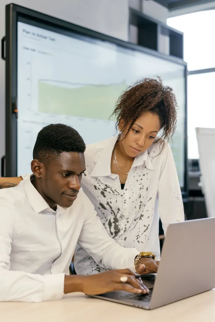 Two colleagues working at a laptop in an office, a dashboard on the screen behind them.